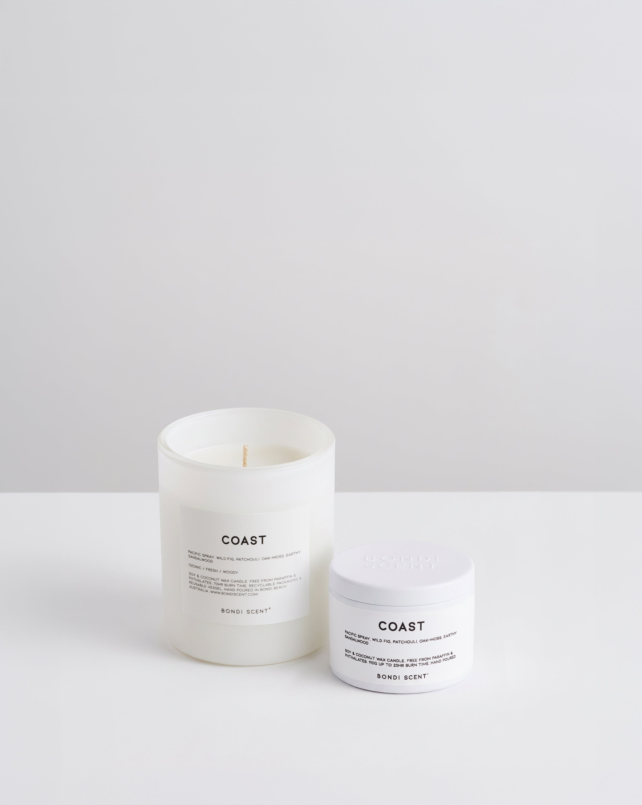 A white glass jar candle called Coast and a white tin candle displayed on a clean white surface against a light gray background.
