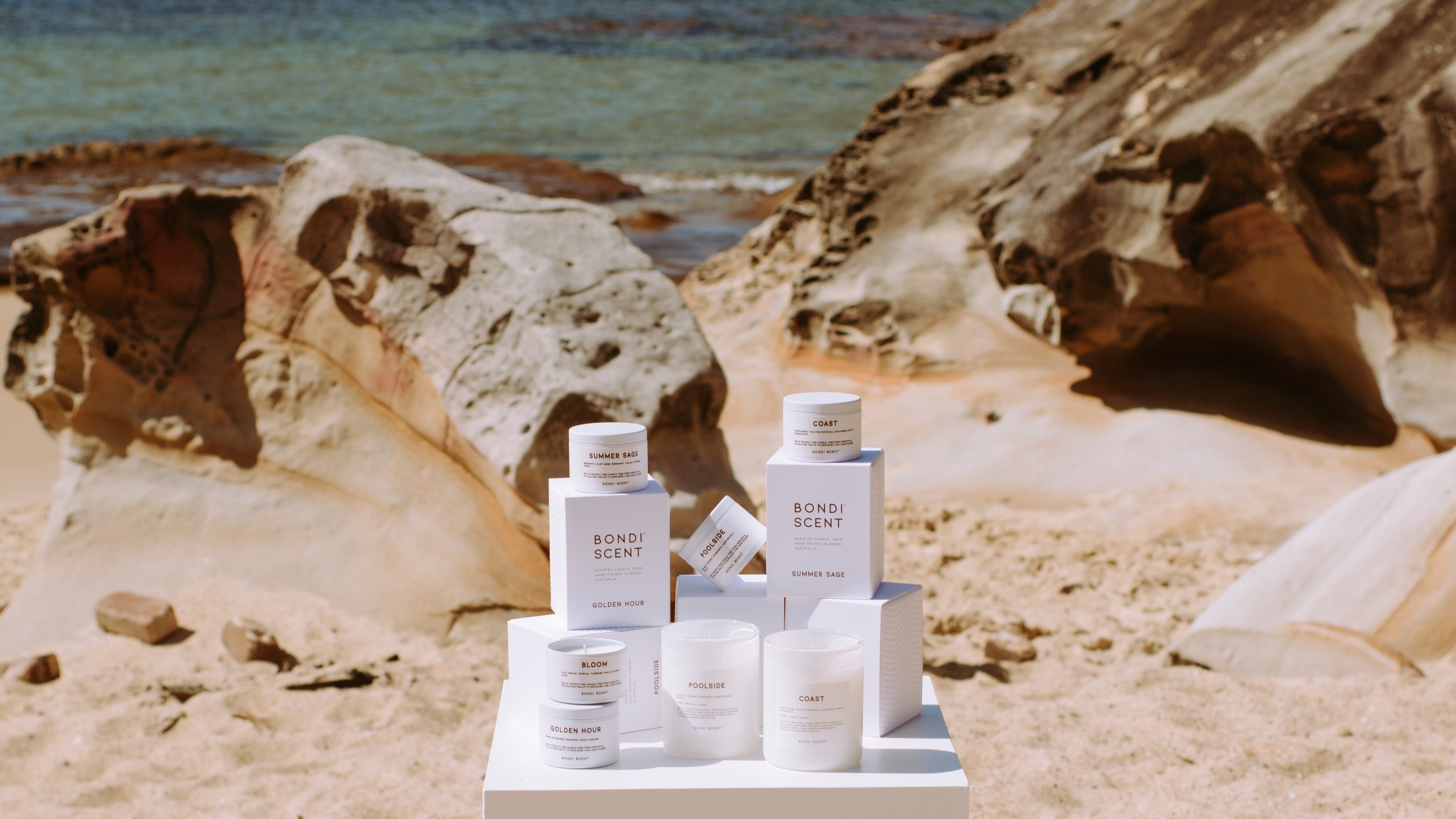 Bondi Scent scented candles and boxes displayed on pedestal by the ocean with rocks and sand