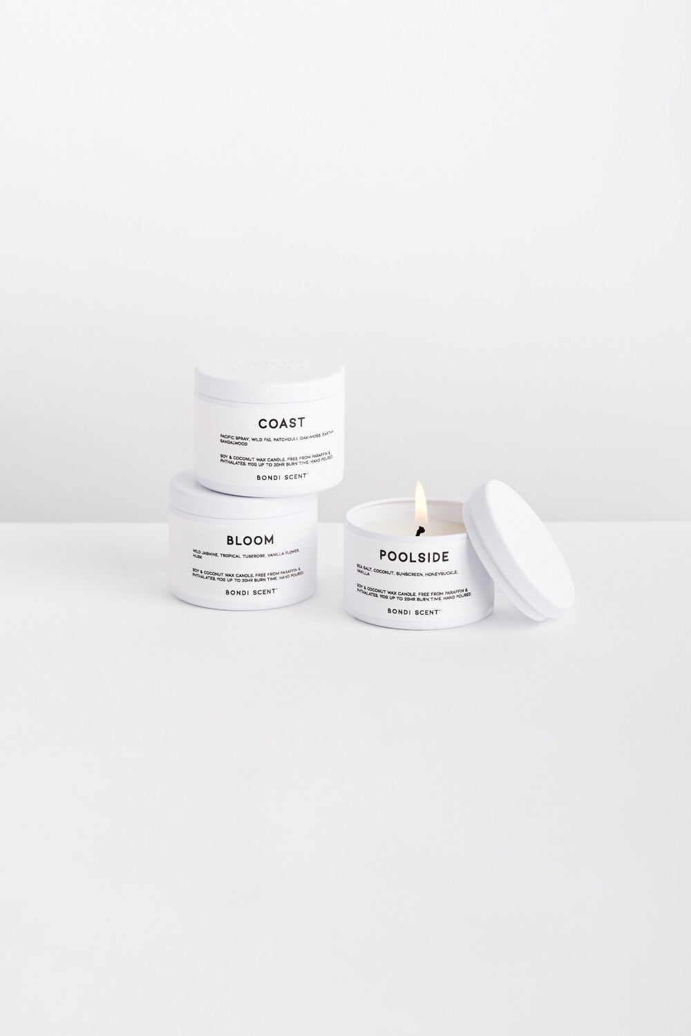 Three white body butter jars with black text labels are arranged on a clean white surface against a white background. One jar is labeled COAST, one labeled BLOOM, and one open jar labeled POOLSIDE with a lit wick and lid resting beside it.