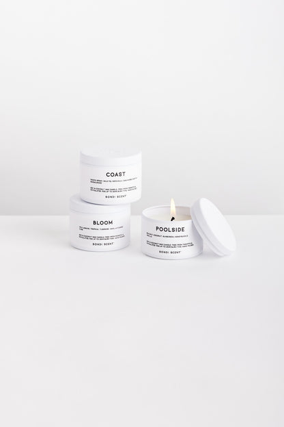 Three white body butter jars with black text labels are arranged on a clean white surface against a white background. One jar is labeled COAST, one labeled BLOOM, and one open jar labeled POOLSIDE with a lit wick and lid resting beside it.