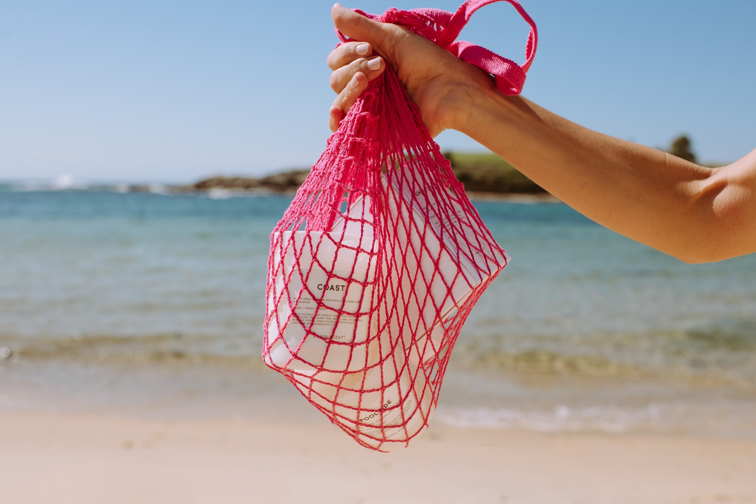 Hand holding a 'Bondi Scent Coast' candle in a pink mesh bag by the ocean.
